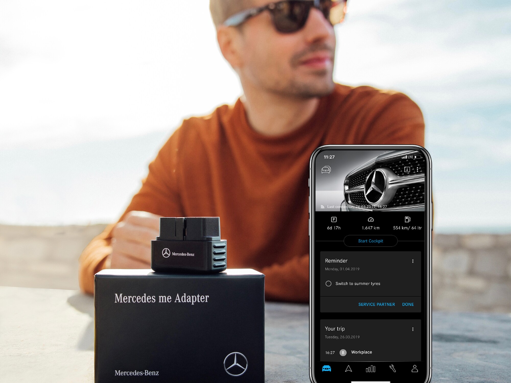 Um homem mostra o Mercedes me Adapter e a Aplicação Mercedes me no smartphone.