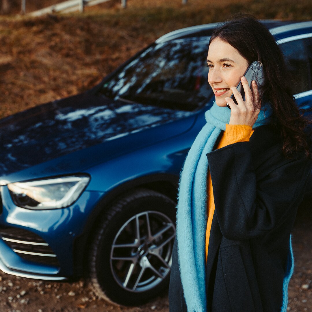 Uma mulher em frente do seu Mercedes SUV a fazer uma chamada com o seu smartphone.