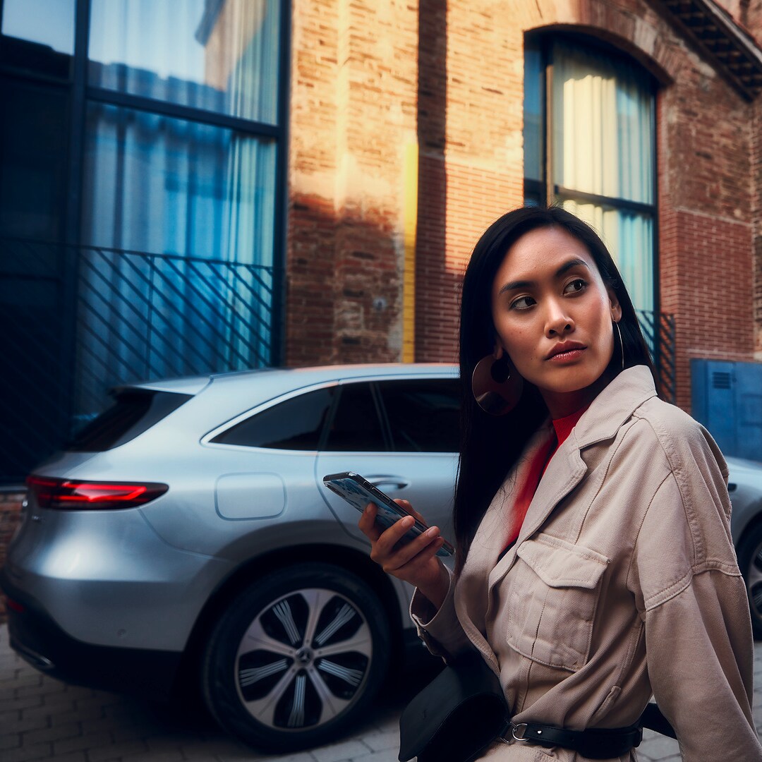 Uma mulher em frente ao seu Mercedes com um smartphone na sua mão direita.
