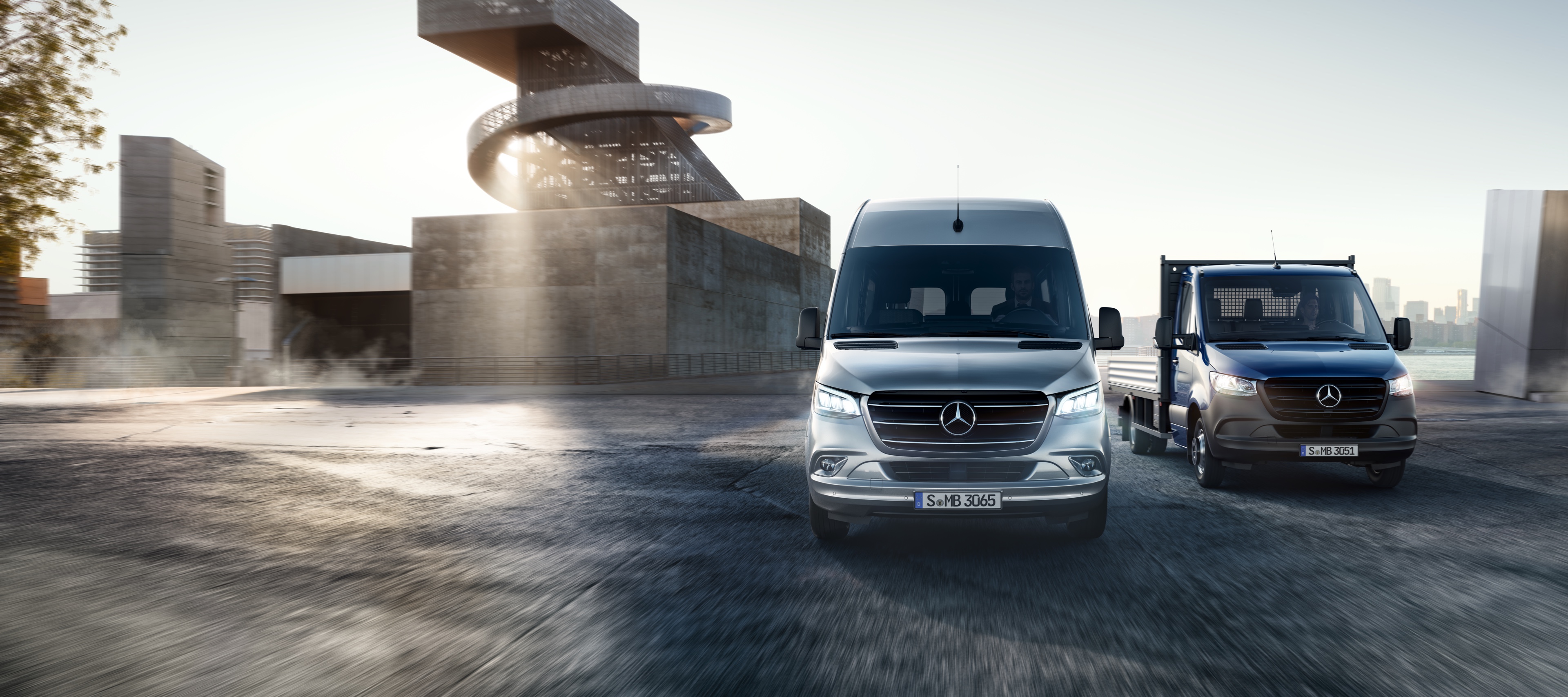 Sprinter | Veículos comerciais ligeiros Mercedes-Benz O Mercedes-Benz Sprinter como carroçaria de plataforma e furgão lado a lado em frente a um conjunto de edifícios modernos.