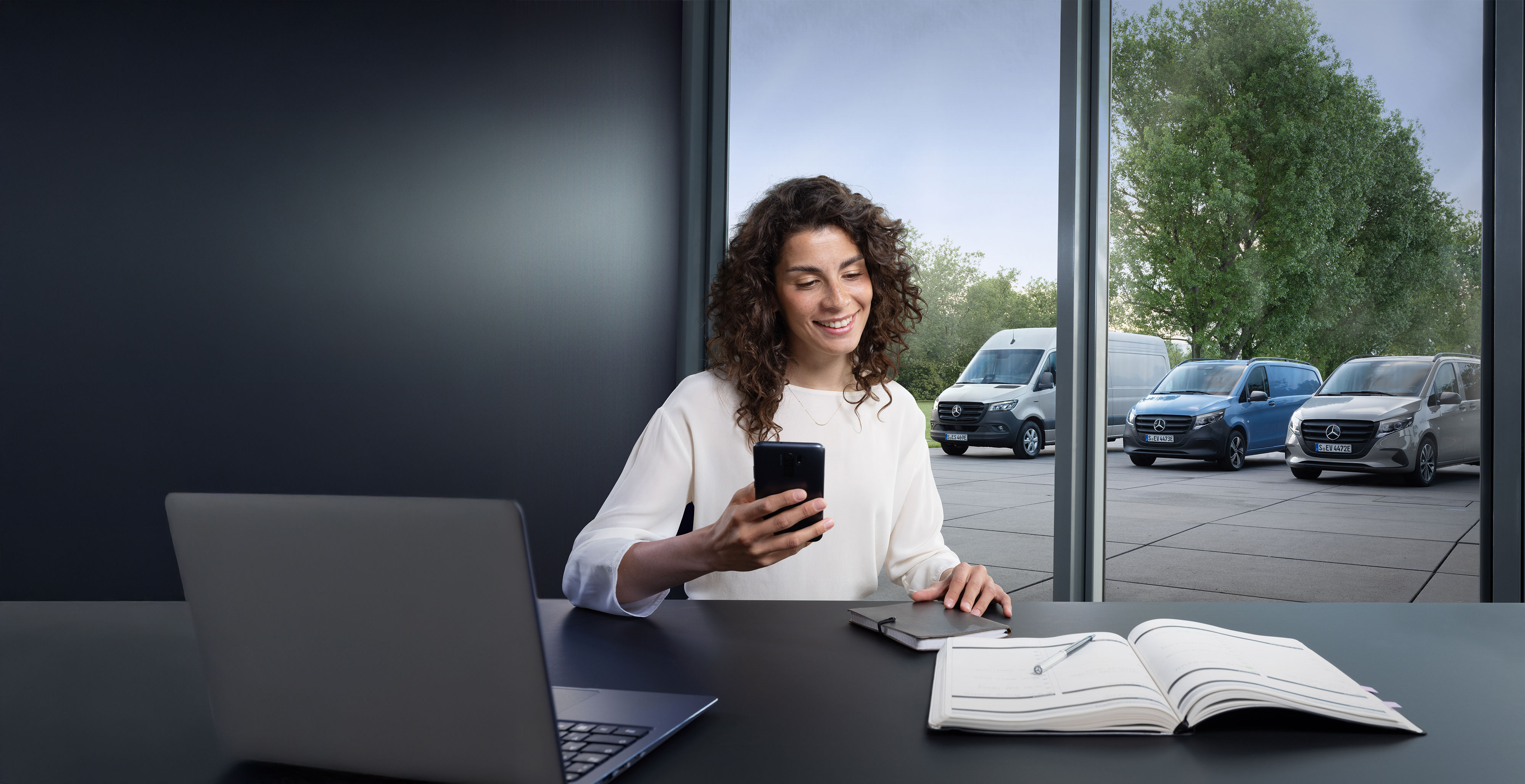 Mercedes-Benz | Assistência de veículos comerciais ligeiros Mulher com smartphone numa secretária, vários veículos comerciais ligeiros Mercedes-Benz em segundo plano: apoio especializado na assistência a profissionais.