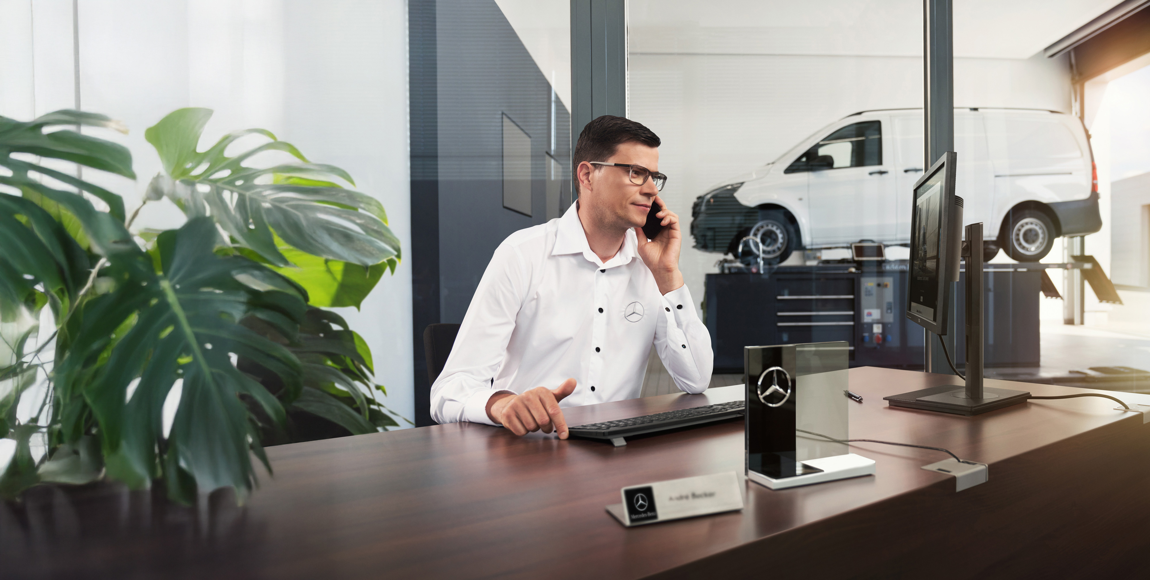 Mercedes-Benz | Assistência de veículos comerciais ligeiros Funcionário da Mercedes-Benz fala ao telefone numa secretária com um veículo comercial ligeiro Mercedes-Benz Vito na oficina.